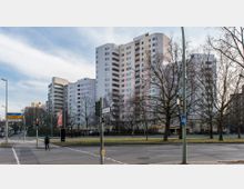 Mehrstöckige Wohngebäude mit heller Fassade und leicht farbigen Akzenten im Stadtgebiet, umgeben von kahlen Bäumen und einer mehrspurigen Straßenkreuzung mit Verkehrs- und Straßenschildern. Im Vordergrund sind ein Fußgängerüberweg, eine Ampel und ein Radweg-Schild zu sehen.