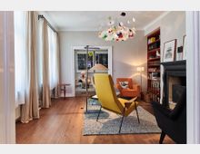 Wohnzimmer mit Holzboden, farbenfrohem Kronleuchter und moderner Einrichtung. Es gibt zwei farbige Sessel (gelb und orange), einen Kamin, ein rotes Bücherregal, große Fenster mit beigefarbenen Vorhängen und einen Teppich unter den Möbeln. Im Durchgang ist ein weiteres Zimmer mit Dekorationen sichtbar.