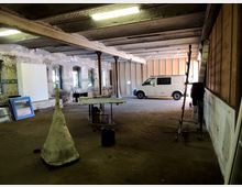 Großer, leerer Industrie-Raum mit sichtbaren Backsteinwänden, Stützpfeilern und einer Betondecke. Im Raum stehen ein weißer Transporter, Arbeitsmaterialien, Leitern und ein tischartiger Aufbau, während an der Wand Fenster und blau gerahmte Gegenstände zu sehen sind.