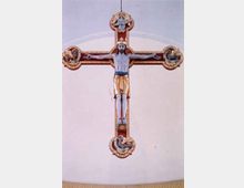 Holzkruzifix mit bemaltem Corpus Christi, umgeben von Verzierungen und vier Evangelistensymbolen an den Enden der Kreuzbalken; Hintergrund ist eine helle, schlichte Wand.