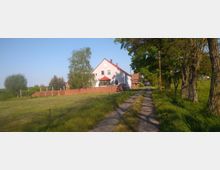 Ein helles, zweistöckiges Wohnhaus mit rotem Satteldach steht hinter einer Mauer aus rotem Backstein, umgeben von Bäumen und Rasenflächen. Rechts führt ein schmaler Pflasterweg, gesäumt von hohen Bäumen, durch eine ländliche Gegend.