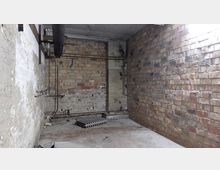 Leerer, heruntergekommener Raum mit unverputzten Ziegelwänden, sichtbaren Rohren und einem Betonboden; auf dem Boden liegt ein Heizkörper.