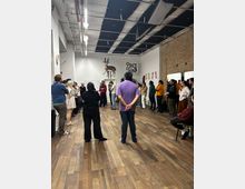 Large open-plan studio or event space with wood plank flooring, white walls, and an exposed brick wall, featuring a grid of ceiling panels with visible pipes and strip lighting. Artwork and signage hang on the walls, including a mounted antelope sculpture above a doorway and a “Price & Son” graphic.