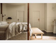 Helles Schlafzimmer mit einem großen Himmelbett, hellem Leinenbettzeug und einer gepolsterten Sitzbank am Fußende. Im Hintergrund sind weiße, holzgetäfelte Wände mit zwei Türen und goldenen Türklinken sowie ein Beistelltisch mit dekorativen Vasen sichtbar.