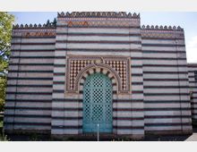 Backsteinfassade eines Gebäudes im maurischen Stil mit klaren horizontalen Streifen in Rot, Weiß und Blau. Zentral ein großes, türkisfarbenes Rundbogenportal mit verzierten Gittermustern und geometrischen Ornamenten im Bogenfeld.