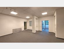 Leerer Büroraum mit hellen Wänden, einem grauen Teppichboden und einer abgehängten Decke mit Einbauleuchten. Der Raum hat mehrere Türen, darunter eine Glastür, die auf einen Balkon oder Außenbereich führt.