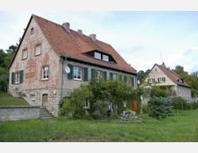 Zwei historische Wohnhäuser mit roten Ziegeldächern und verputzten Fassaden, teils mit Efeubewuchs. Vor den Häusern befindet sich ein Gartenbereich mit Rasen, Bäumen und Sitzgelegenheiten.