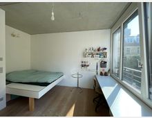 Minimalistisch eingerichtetes Zimmer mit Beton- und Holzelementen, einem schwebenden Bett links, einem langen weißen Schreibtisch vor einem großen Fenster rechts und Regale sowie Wandpaneele mit kleinen Pflanzen und Schreibutensilien an der Wand.