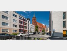 Straßenszene mit Wohngebäuden auf beiden Seiten und einer Backsteinkirche mit spitzem Turm und roten Dachziegeln im Hintergrund. Geparkte Autos stehen entlang der Straße, die unter einem blauen Himmel mit wenigen Wolken verläuft.