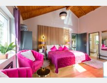 Ein gemütliches Schlafzimmer mit Holzbalkendecke, einem Doppelbett mit pinker Bettwäsche und passenden Kissen sowie zwei pinken Sesseln. Im Raum befinden sich ein Fenster mit Vorhängen, ein Nachttisch mit Lampe, eine Holzvertäfelung hinter dem Bett und ein offenes Badezimmer im Hintergrund.