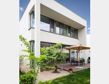 Moderne zweistöckige Wohnung mit einer hellen, kubischen Fassade und großen Fenstern. Vor dem Gebäude befindet sich eine Holzterrasse mit einem Esstisch, einem Sonnenschirm, Pflanzen und einem kleinen Kiesbereich im Garten.