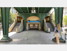 Ein U-Bahnhof-Eingang der Station „Eberswalder Straße“ unter einer grünen Stahlkonstruktion der Hochbahn in Berlin. Treppen führen beidseitig hinauf, und ein überdachter Gang verläuft mittig unter der Stahlbrücke hindurch. Eine Person geht auf die Szene zu, und ein Elektroroller steht seitlich geparkt.