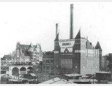 Historische Aufnahme eines Kühlhauses mit Ziegelfassade und Türmen, beschriftet mit „Gesellschaft für Markt-Kühlhallen, Kühlhaus I“. Im Vordergrund sind ein Viadukt, Marktstände und Baugerüste zu sehen.