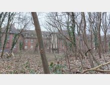 Backsteingebäude mit Satteldach und Dachfenstern, teilweise von kahlen Bäumen und dichter Vegetation eines Waldes verdeckt. Der Boden ist mit Laub bedeckt, und das Gebäude wirkt verlassen.