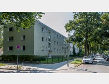 Mehrstöckiges Wohngebäude mit grün gestrichener Fassade und mehreren Fenstern, umgeben von einem kleinen Vorgarten mit Zaun. Rechts im Bild verläuft eine Baumallee mit parkenden Autos entlang einer gepflasterten Straße, ein Straßenschild weist auf die Brahmsstraße hin.