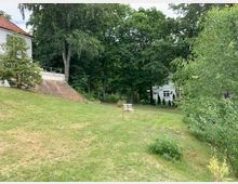 Grüne Gartenlandschaft mit einer Rasenfläche, Bäumen und Blumenbeeten; links ein Teil eines Gebäudes mit weißer Fassade und rotem Dach, im Hintergrund ein weiteres weißes Gebäude hinter den Bäumen. Im Vordergrund stehen zwei leere Gartenstühle.
