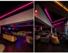 Innenraum einer modernen Lounge- oder Bar mit niedriger Decke und farbigen Neon-Lichtleisten in Pink, Blau und Orange. Samtsofas und gepolsterte Sessel gruppieren sich um niedrige Tische, im Hintergrund sind eine Bar und ein abgetrennter Bühnen- oder Vorhangbereich zu sehen.