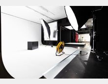 Professionelles Fotostudio mit weißem Hintergrund, großen Softboxen, Reflektoren und einem Ventilator auf einem weißen Boden. Im Hintergrund sind weitere Sets mit schwarzem und rotem Stoff zu sehen.