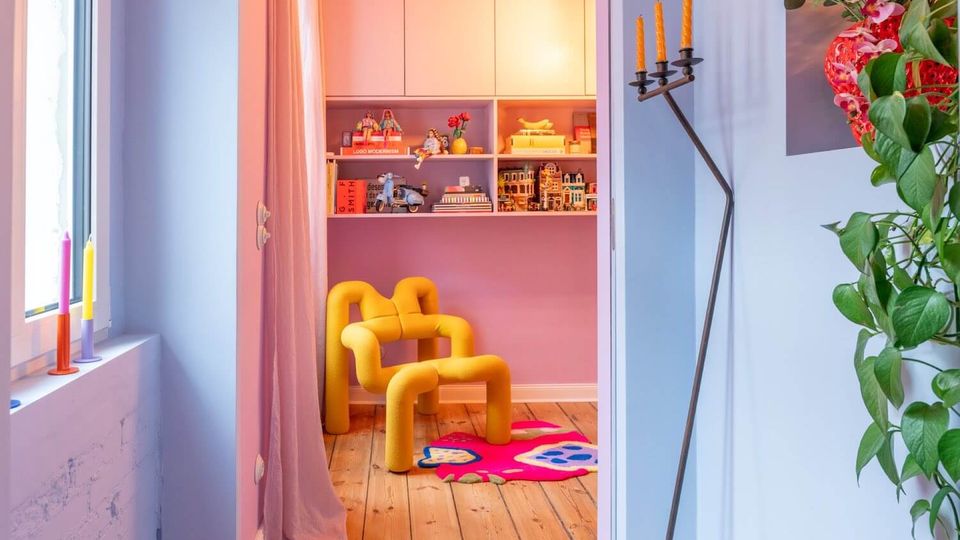 Cozy, colorful reading nook with a bright yellow sculptural lounge chair on a wooden floor, set in front of built-in shelves filled with books and small figurines. A window with sheer curtains, candle holders, and a tall minimalist floor candelabra frame the space, with a pink wall and patterned rug adding color.