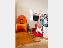 Ein Kinderzimmer mit Holzboden: Links steht ein orangefarbener Stuhl mit einer Spielzeugfigur darauf, daneben ein Beistelltisch mit einer weißen Skulptur eines Vogels und einem kleinen Verstärker. Rechts lehnt eine rote E-Gitarre, und ein Bett mit bunter Bettwäsche ist teilweise sichtbar. Ein Fellteppich liegt teilweise auf dem Boden.