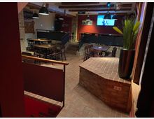 Barraum mit rustikalem Design, sichtbar durch Backsteinwände, rotbraune Fliesen und Holztische mit dunklen Stühlen. Im Hintergrund befinden sich eine Projektionsfläche, schwarze Pendelleuchten und eine erhöhte Sitzfläche, dekoriert mit schlichten Akzenten.