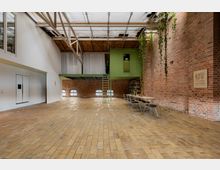 Industrieller Innenraum mit Ziegelwänden, großen Fenstern und einem offenen Raum mit gelbem Backsteinboden. Eine grüne Zwischendecke verbindet den oberen Bereich, mit einer Leiter als Zugang; ein langer Holztisch mit Stühlen steht an der Wand, über die Pflanzen von der Decke hängen.