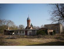 Verlassener Gebäudekomplex mit niedrigen, teils verfallenen Nebengebäuden vor einem hohen Backsteinturm mit Metallkuppel. Davor liegt eine überwachsene Wiese, umgeben von kahlen Bäumen und weiteren alten Mauerresten unter klarem Himmel.