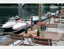 Ein Außenbereich eines Restaurants oder Cafés direkt an einem Fluss mit Holzstühlen und Tischen, auf denen Essen, Bierflaschen und Blumen in einer Vase stehen. Im Hintergrund liegen kleine motorisierte Boote am Steg, und eine Brücke ist sichtbar.