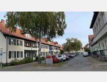 Straßenszene in einer Wohngegend mit zweigeschossigen Häusern, die helle Fassaden und rote Ziegeldächer haben. Links ist ein schmaler Gehweg mit Bäumen, rechts parkende Autos und weitere Wohnhäuser mit großen Fenstern.