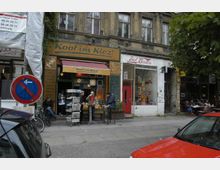 Straßenszene mit einem kleinen Laden namens „Koof im Kiez!“ in einem alten mehrstöckigen Gebäude mit verwitterter Fassade. Vor dem Laden stehen Verkaufsständer mit Zeitungen sowie Personen an Stehtischen, daneben ein Geschäft mit roter Tür und bunter Dekoration im Fenster.