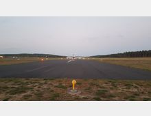 Ein kleiner Flugplatz mit einer asphaltierten Landebahn, auf der ein Flugzeug am Ende der Bahn steht. Die Umgebung ist flach und von Wiesen und Wäldern begrenzt, mit vereinzelten Signallichtern entlang der Piste.