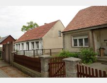 Zwei kleine Einfamilienhäuser mit Satteldächern und roten Dachziegeln, getrennt durch einen schmalen Gartenzaun. Vor den Häusern befinden sich Vorgärten mit Blumen und Sträuchern, eingefasst von Zäunen aus Holz und Metall.