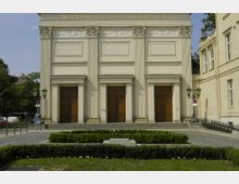 Fassade des Maxim-Gorki-Theaters in Berlin mit klassischen architektonischen Elementen wie Säulen, großen Holztüren und kunstvollen Reliefs oberhalb der Türen. Im Vordergrund ein kleiner gepflasterter Platz mit niedrigen Buchsbaumhecken und Laternen.