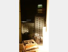 Nächtliche Ansicht eines beleuchteten historischen Gebäudes mit mehreren Fenstern und einem Turm mit gläserner Kuppel, gesehen durch ein Fenster. Im Vordergrund befinden sich gestapelte CDs und eine leuchtende Tischlampe.