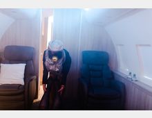 Eine Flugzeugkabine mit zwei Ledersesseln, umgeben von Holzwänden. Im Vordergrund beugt sich eine Person in Schutzkleidung mit Atemmaske nach vorne, während Rauch den Raum leicht einhüllt.