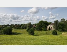 Weite Wiesenlandschaft mit gelbem Blumenbewuchs, umgeben von Bäumen und vereinzelten Gebäuden, darunter ein großes, helles Haus mit dunkelgrauem Dach. Der Himmel ist mit vielen Wolken bedeckt.