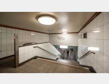 Treppenabgang in einer U-Bahn- oder S-Bahn-Station mit weißen, gefliesten Wänden und Handläufen. Auf der Wand befindet sich eine Beschilderung mit den Linien "S1 Wannsee", "S2 Blankenfelde" und "S25 Teltow Stadt".