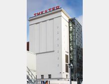 Außenansicht eines weißen Theatergebäudes mit glatter Fassade und rotem „THEATER“-Schriftzug auf dem Dach. Rechts schließt ein moderner, verglaster Anbau mit sichtbarem Treppenhaus und Metallrahmen an.