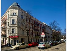 Ecke eines historischen Wohnhauses mit verzierten Fassaden in Weiß und Rot in einer städtischen Straße, vor der ein Café mit der Aufschrift „Café Kurve“ liegt. Parkende Autos entlang des Gehwegs und Bäume ohne Blätter rahmen die Straßenszene.