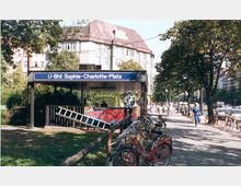 Außenansicht des U-Bahnhofs Sophie-Charlotte-Platz in Berlin mit einem überdachten Eingangsbereich und einem blauen Schild mit der Stationsbezeichnung. Im Vordergrund stehen Fahrräder an einem Zaun, im Hintergrund sind Bäume sowie ein historisches Gebäude mit rotem Dach zu sehen.