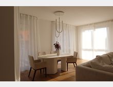 Heller, modern eingerichteter Wohn- und Essbereich mit einem ovalen weißen Esstisch, vier gepolsterten Stühlen und einem Strauß Blumen als Dekoration. Große Fenster mit lichtdurchlässigen Vorhängen sorgen für viel Tageslicht; ein Teil eines Sofas ist im Vordergrund sichtbar.