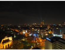 Nachtaufnahme einer Stadt mit beleuchteten Straßen und Gebäuden. Im Vordergrund ist ein historischer Torbogen erleuchtet, während im Hintergrund moderne Gebäude und grelle Lichter die Skyline prägen.