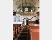 Innenraum einer kleinen Kirche mit Holzbänken in rötlichem Farbton, einem Mittelgang mit Teppich, weißer Orgel an der linken Wand und einer halbkreisförmigen Empore. Die Decke ist gewölbt, und eine geschmückte Hängelampe ist zentral positioniert; eine offene Holztür führt nach außen.