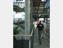 Ein modernes Gebäude mit einer offenen, industriellen Architektur, gekennzeichnet durch Metallgeländer, sichtbare Stahlstrukturen und eine Betonsäule. Treppen und Galerien mit Holz- und Metallelementen sind zu sehen, während im Vordergrund zwei Personen einen Gang entlanggehen; im Hintergrund stehen Glaswände und Pflanzen.