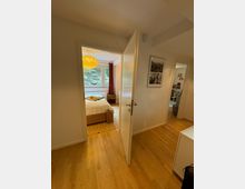 Blick durch eine geöffnete Tür in ein Schlafzimmer mit einem Holzboden, einem Bett mit beigefarbener Tagesdecke und einem Korb am Fußende. Das Zimmer hat ein großes Fenster mit Blick auf eine Mauer aus Natursteinen und ist beleuchtet von einer hängenden, runden Lampe.