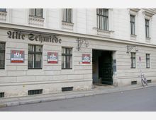 Fassade eines historischen Gebäudes mit der Aufschrift „Alte Schmiede“ und Schildern, die „Zur Alten Schmiede – Lokal“ bewerben. Rechts befindet sich eine geöffnete, dunkle Holztür, die in einen Hofeingang führt; daneben lehnt ein Fahrrad an einem Verkehrsschild.