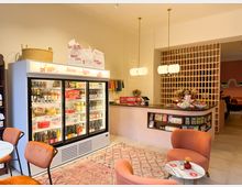 Ein kleiner, hell beleuchteter Laden mit einer Kühltheke voller Getränke und Lebensmittel, einem Verkaufstresen mit dekorativen Lampen und Regalen mit Süßigkeiten. Im Vordergrund stehen orangefarbene Stühle und ein runder Marmortisch auf einem gemusterten Teppich.