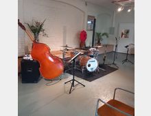 Ein Musikprobenraum mit hellem Holzboden und weißer Ziegelwand. Zu sehen sind ein Kontrabass, ein Schlagzeugset, ein Saxophon, Notenständer und Mikrofone, sowie Dekorationselemente wie Pflanzen und ein großes rotes Kunstobjekt.