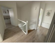 Heller Flur in einem modernen Wohnhaus mit Laminatboden, einer weißen Treppe mit Glasgeländer nach unten, zwei angrenzenden Räumen mit offenen Türen und minimalistischer Wanddekoration.