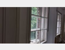 Blick in einen Raum mit großen, weißen Holzfenstern, die nach außen hin auf üppige grüne Vegetation und eine Ziegelwand zeigen. Der Innenraum wirkt verwittert, mit abblätternder Farbe und sichtbaren Gebrauchsspuren.